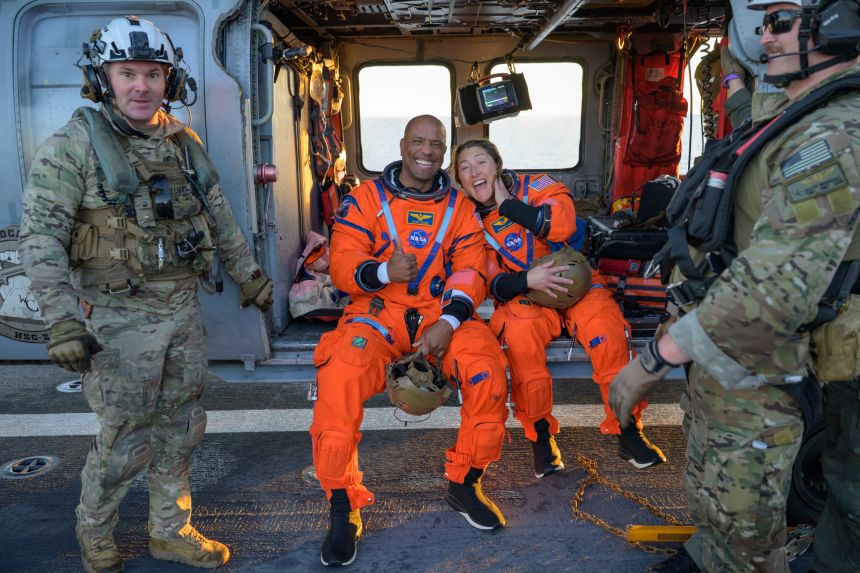 Victor Glover de la NASA (al doilea de la stânga) și Christina Koch stau pe un Navy MH-60 Seahawk pe puntea de zbor a USS John P. Murtha, după stropire de vineri.